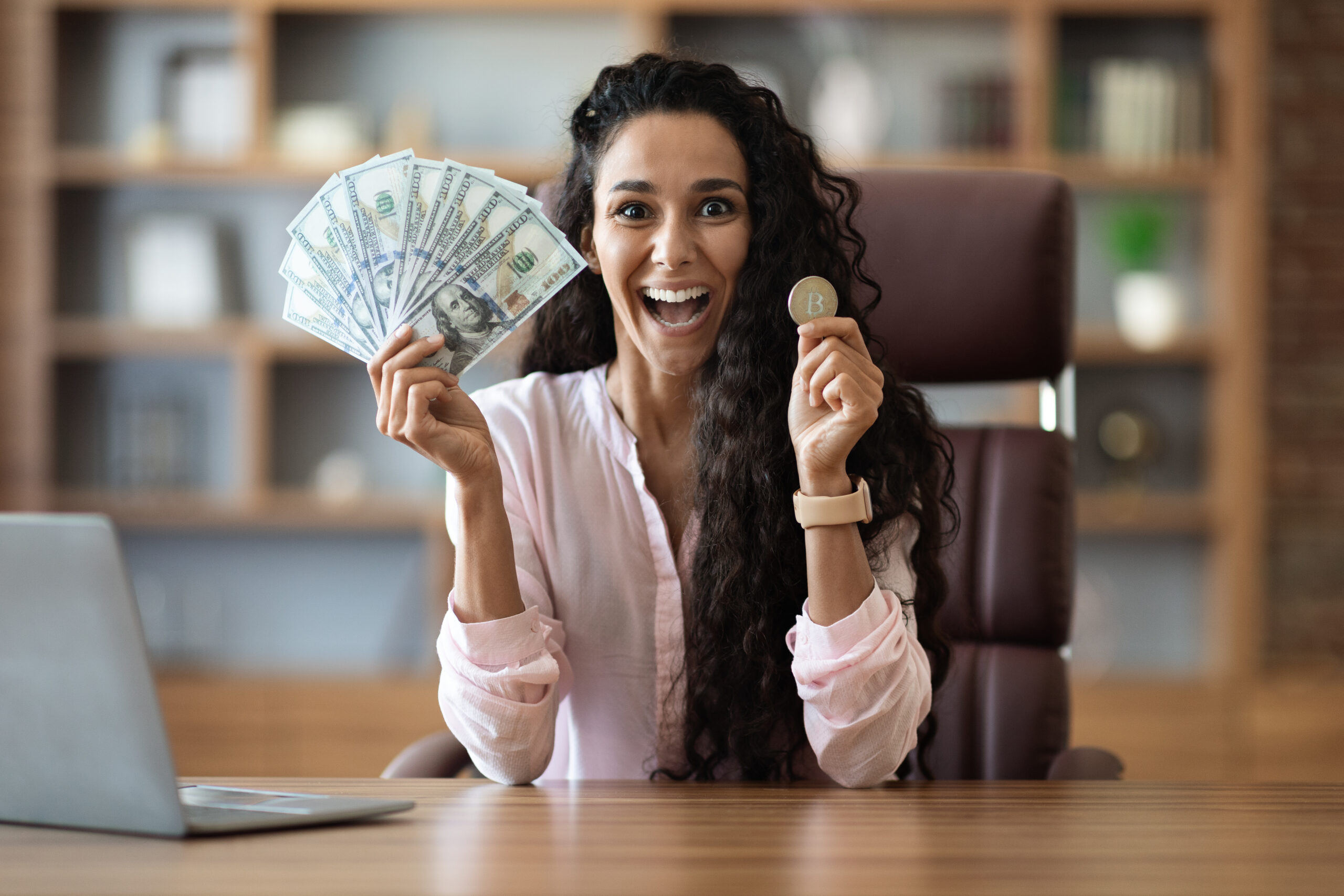 Happy businesswoman holding cash and bitcoin, office interior