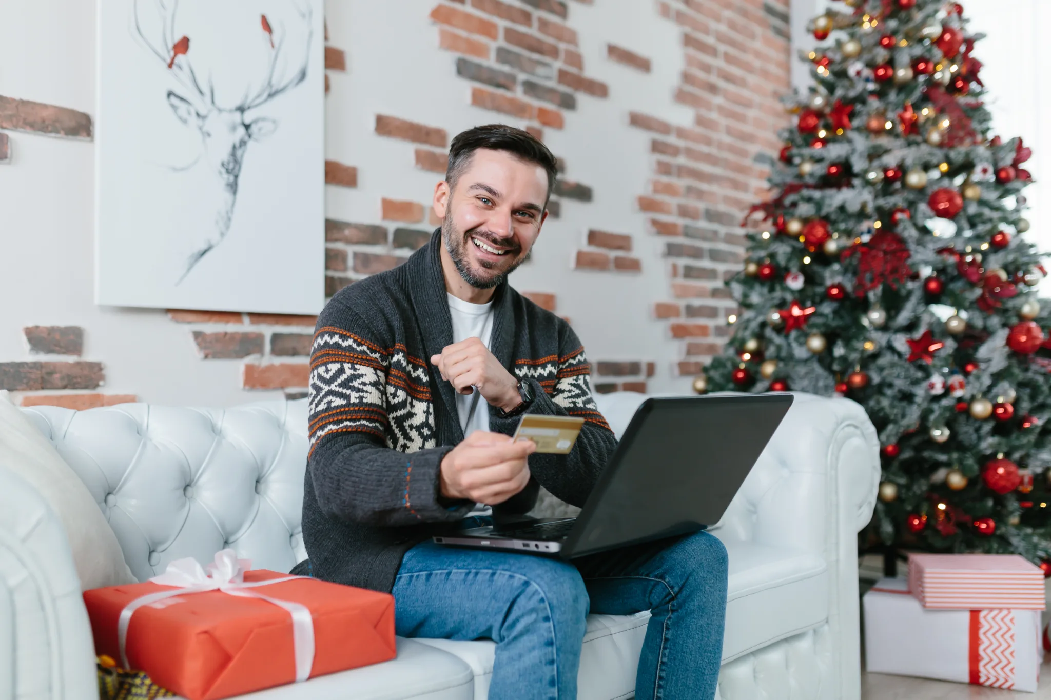 Playful man with a beard smiling sitting on the couch shopping online, buying Christmas presents, online store using laptop