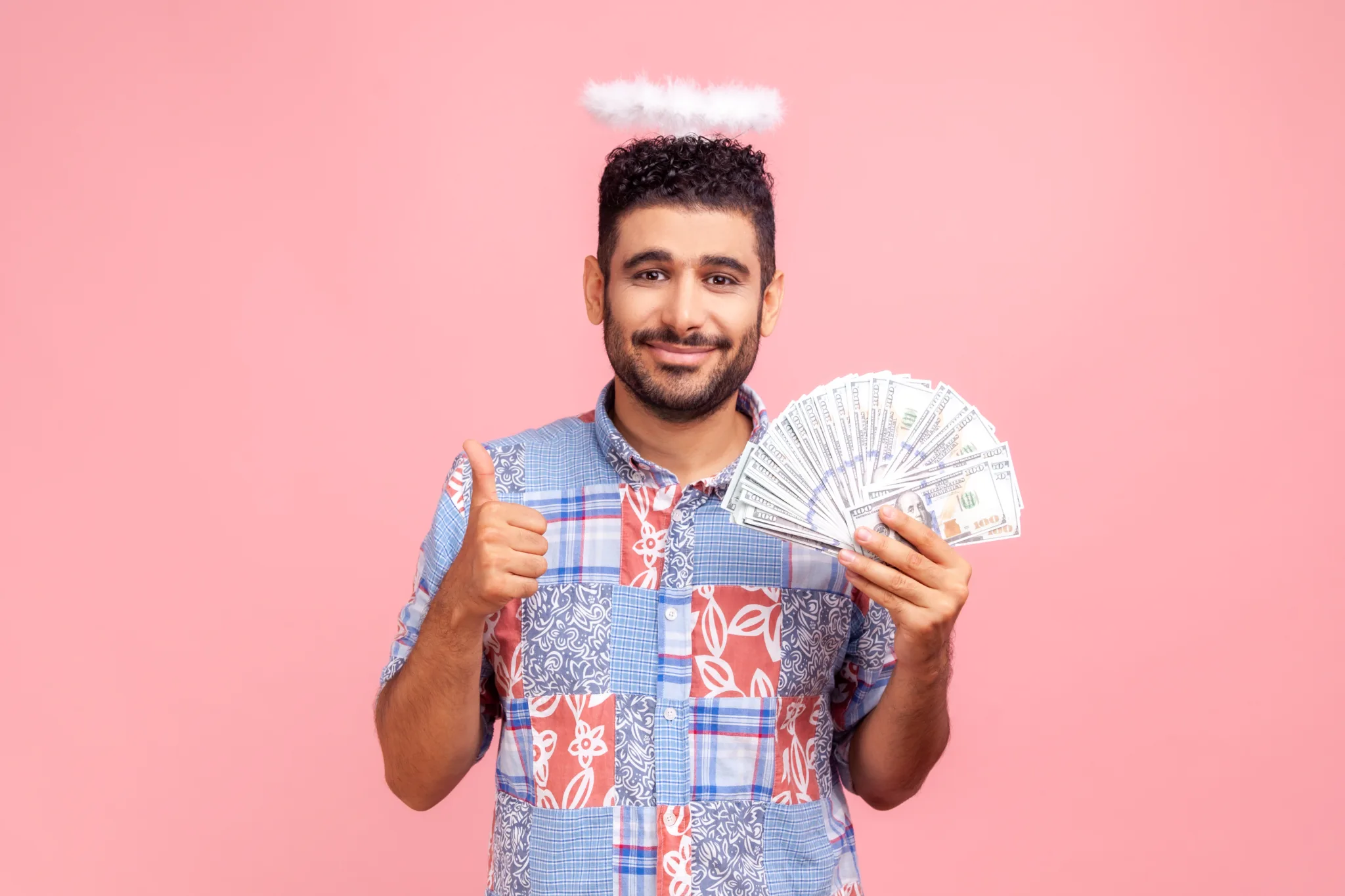 Smiling positive man with beard wearing blue casual style shirt with nimbus on head holding dollars banknotes and showing thumb up.
