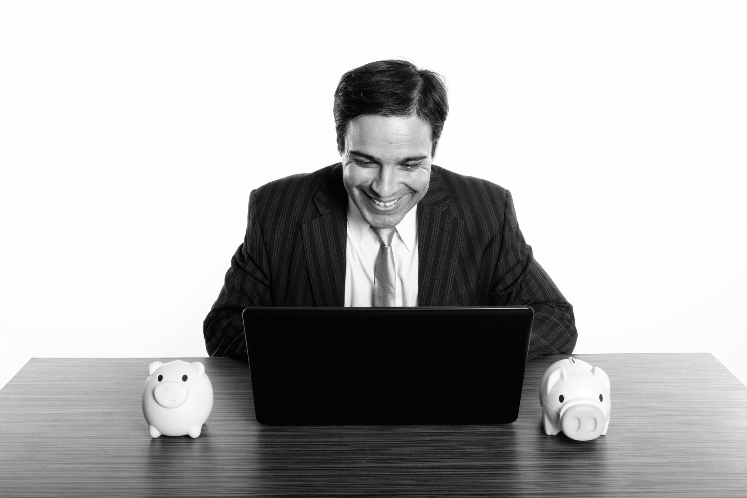 Young happy Persian businessman smiling while using laptop with two piggy banks on wooden table